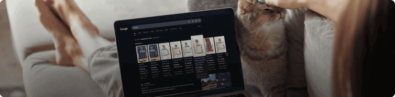 A lady laying on her couch while browsing Google, looking for affordable cat food while scratching her cat affectionately.